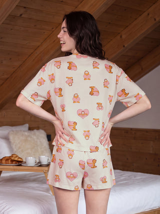 A woman in a cream-colored Garfield print pajama set stands with her back to the camera, a breakfast tray on the bed beside her.