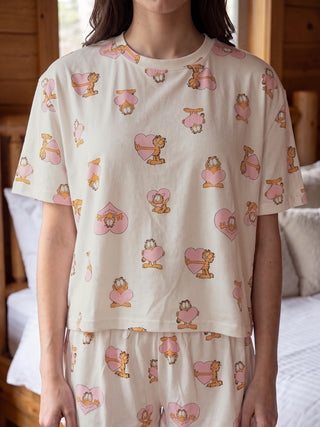 A woman stands wearing a cream-colored Garfield-print pajama set with Garfield characters holding pink hearts, a slightly different print pattern than previous images.