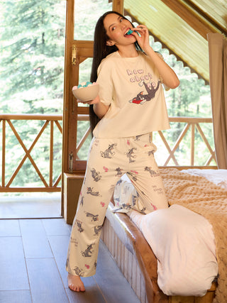 A woman in cream-colored pajamas with Tom and Jerry prints is posing while holding a bowl of candies and eating a gummy.