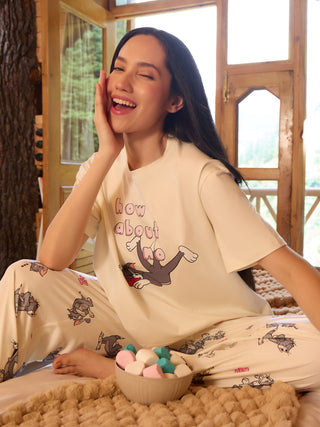 Woman in a cream pajama set with a Tom and Jerry print, winking and holding her cheek as she sits on a bed.