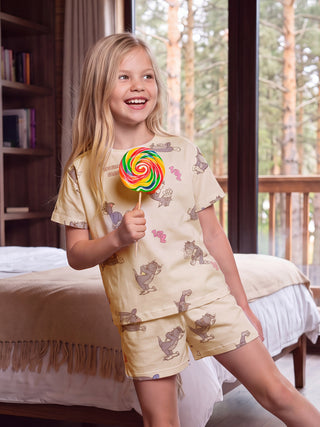 A young girl with blonde hair smiles while holding a colorful lollipop and wearing a light yellow sleepwear set with a cartoon cat print.