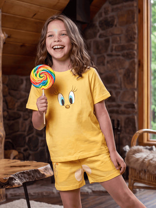 Girls in a yellow Tweety Bird pajama set, featuring a character print on the top and shorts, holding a rainbow lollipop.