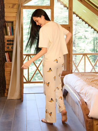 A woman in a cream-colored short-sleeved top and cream-colored pajama pants with a cartoon duck print walks on a wooden floor near a bed.