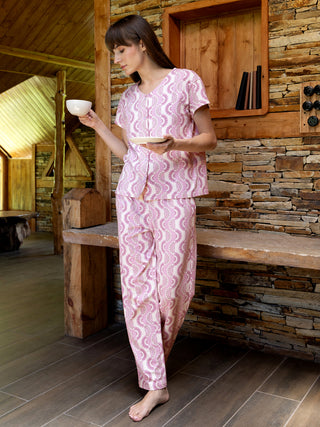A woman stands holding a teacup and saucer, wearing pink and white patterned pajamas.