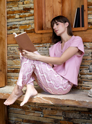 A woman sits cross-legged reading a book, wearing pink pajamas with a white paisley print.