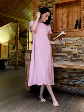 A woman stands on a wooden floor wearing a pink, knee-length nightgown with a bird print.
