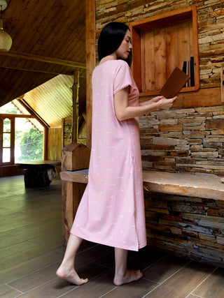 A woman stands with her side to the camera, wearing a light pink nightgown with a delicate white bird print.
