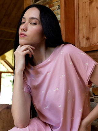 A woman with long black hair poses with her hand on her chin, wearing a pink pajama set with a bird print and lace trim.