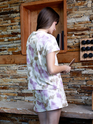 A young woman stands facing left, wearing a tie-dye pajama set with a white, purple, and brown floral print.