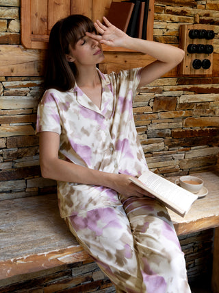 Women's pastel tie-dye pajama set, featuring a collared button-down top and matching pants, with the model sitting with a book and a cup of tea.