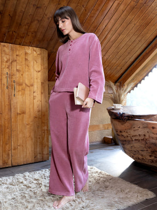 A young woman with bangs stands in a cozy room, wearing a pink terry cloth pajama set.