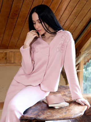 A woman in a pink pajama set with embroidered details sits on a wooden table, resting her chin on her hand.