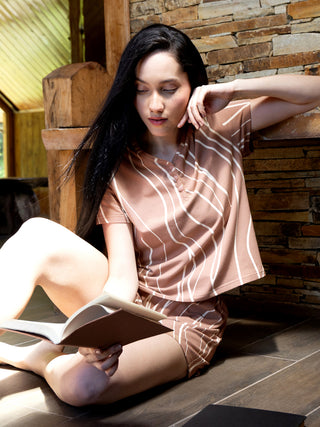 A woman in a brown and white striped pajama set is sitting on the floor and reading a book.