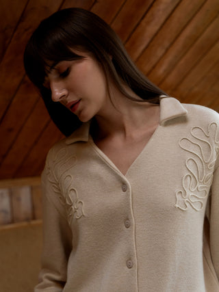 A woman wears a light beige, collared cardigan with embroidered leaf patterns, unlike the previous image of a woman in a similar cardigan, this one is buttoned up.