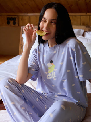 A woman in light purple cloud-print pajamas sits on a bed and eats an apple.