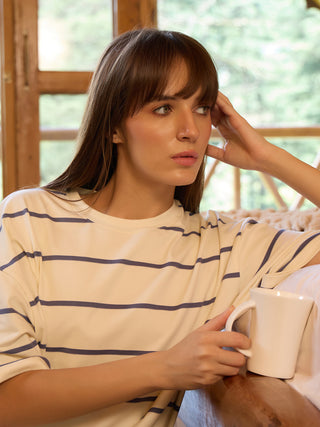 A woman leans her head on her hand while holding a mug, wearing a cream and navy striped oversized t-shirt.
