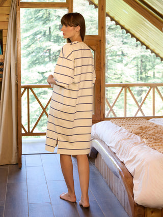 Woman in a cream and navy striped loungewear dress stands facing away from the camera, looking out a window, with a cozy bedroom to her right.