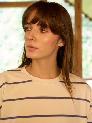 A woman with long brown hair and bangs, wearing a cream-colored sweatshirt with navy blue stripes, is pictured in a close-up shot.