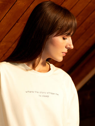 Woman in a cream-colored crewneck sweatshirt with a delicate "where the stars whisper me to sleep" print, shown in a profile shot looking down.