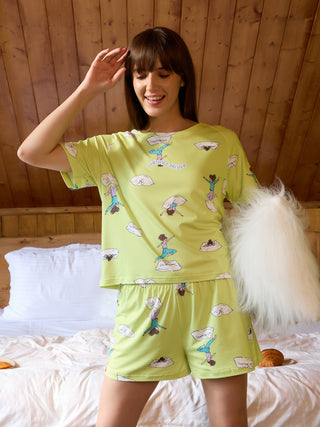 Women in light green pajamas with a "Certified Delulu" print, featuring cartoon figures in yoga poses, stand on a bed.