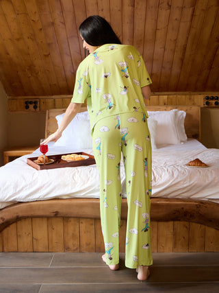 A woman in light green pajamas with a cartoon print is posing by a bed.