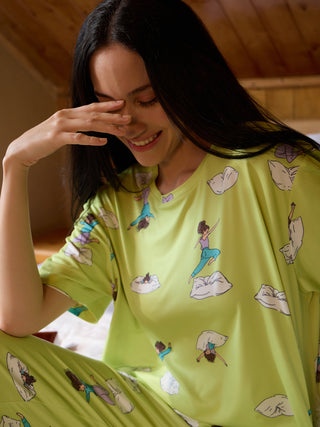 A woman in light green pajamas with a cartoon print of figures in various poses is laughing and covering her eyes.