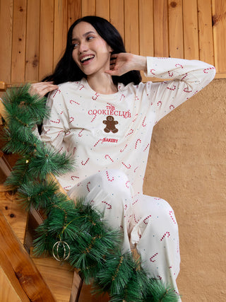 A woman wears cream-colored pajamas with a gingerbread man and candy cane print, sitting on wooden stairs.