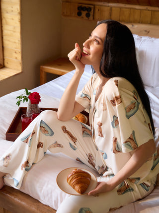 A woman in cream pajamas with a playful dog print, resting on a bed with a croissant and juice.