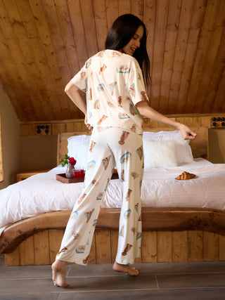A woman in white pajamas with a playful puppy print poses on a bed, with a croissant on the side.