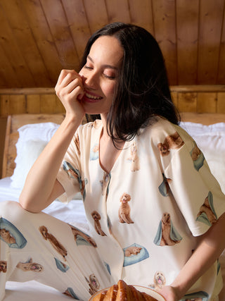 A woman in cream-colored pajamas with a playful puppy print poses on a bed, holding a croissant.