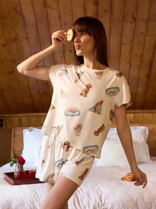 A woman poses with a slice of apple on her eye, wearing cream-colored pajama shorts and a matching t-shirt with a playful dog print.
