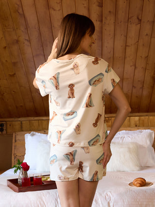 A woman in a cream-colored pajama set with a playful dog print poses with her back to the camera, showcasing the short-sleeved top and matching shorts.