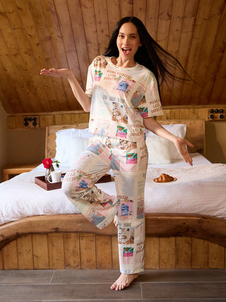 A woman in light-colored pajamas with a newspaper print stands on a bed, posing with one leg bent and her arms outstretched.