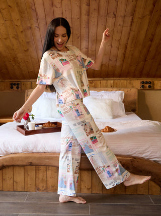 Women in cream pajamas with a newspaper print dance joyfully near a bed.