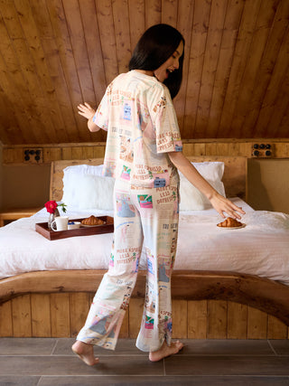 A woman in a light-colored pajama set with a newspaper-like print featuring playful illustrations and text, poses playfully on a bed.