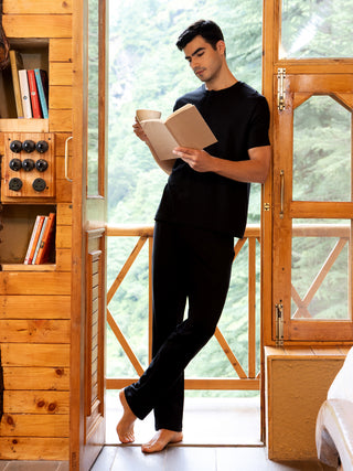 A man in black sleepwear is reading a book and holding a cup, leaning against a wooden doorframe with a backdrop of lush green trees.