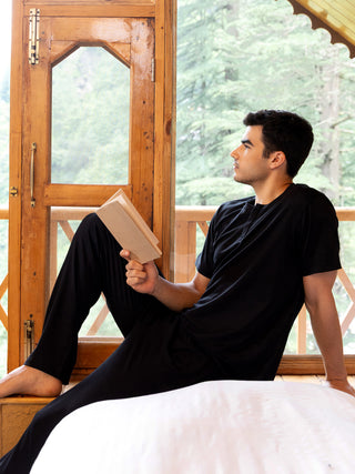 Men in black loungewear reading a book by the window.