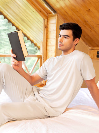 A young man sits on a bed, wearing a light grey sleepwear set, holding a book and looking away from the camera.