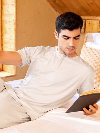 A man in light gray sleepwear lounges on a bed, reading a book.