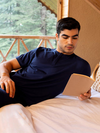 A man in a navy blue sleep shirt reclines on a bed reading a book.