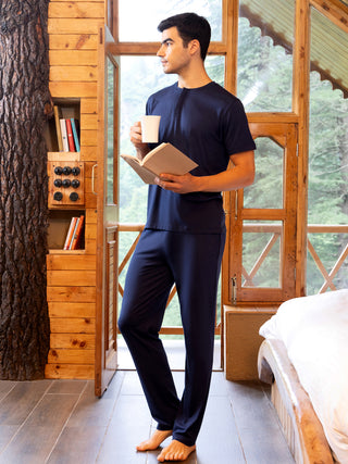 A man in dark blue pajamas holding a book and a mug, standing in a rustic cabin room.