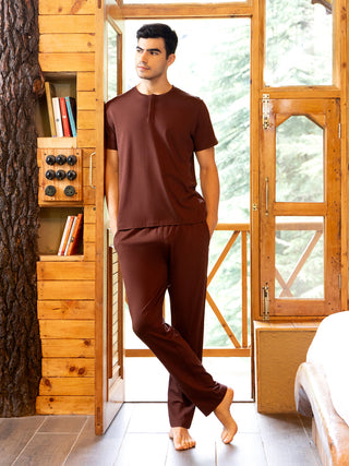 A man poses wearing a brown henley sleep shirt and sleep pants.