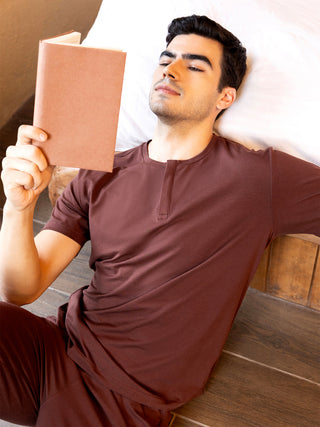Men's maroon short-sleeve Henley sleepwear top and pants set, posed reading a book in bed.