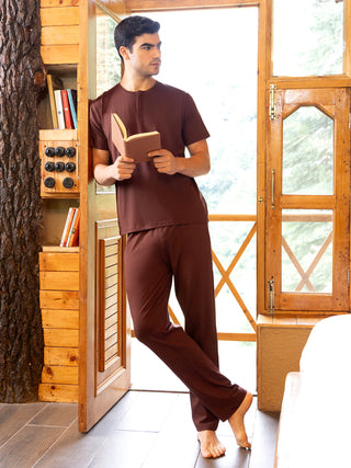 Men in brown sleepwear, holding a book, leaning against a wooden doorframe.