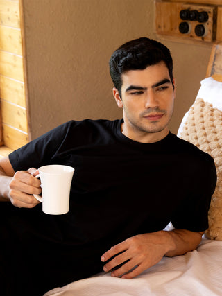 A man in a black t-shirt holds a white mug while resting in bed.