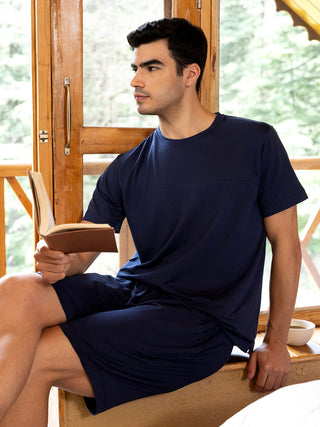 Men wearing navy blue short-sleeved pajamas, sitting with a book, with a cup of coffee nearby.