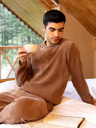 A man in a brown corduroy pajama set is sitting on a bed, holding a cup of coffee and looking down at an open book.