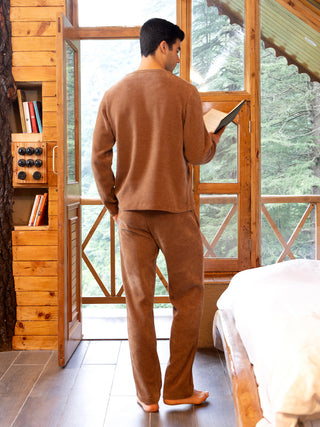 A man in a brown loungewear set stands by a window reading a book.