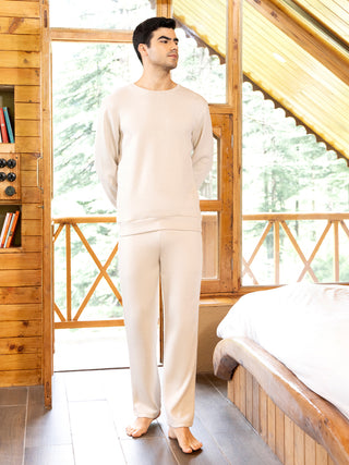 Men in a cream-colored loungewear set standing barefoot next to a bed with a window view.