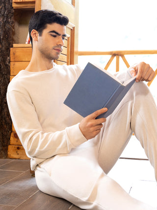 A man in cream-colored loungewear is sitting on the floor, reading a book.